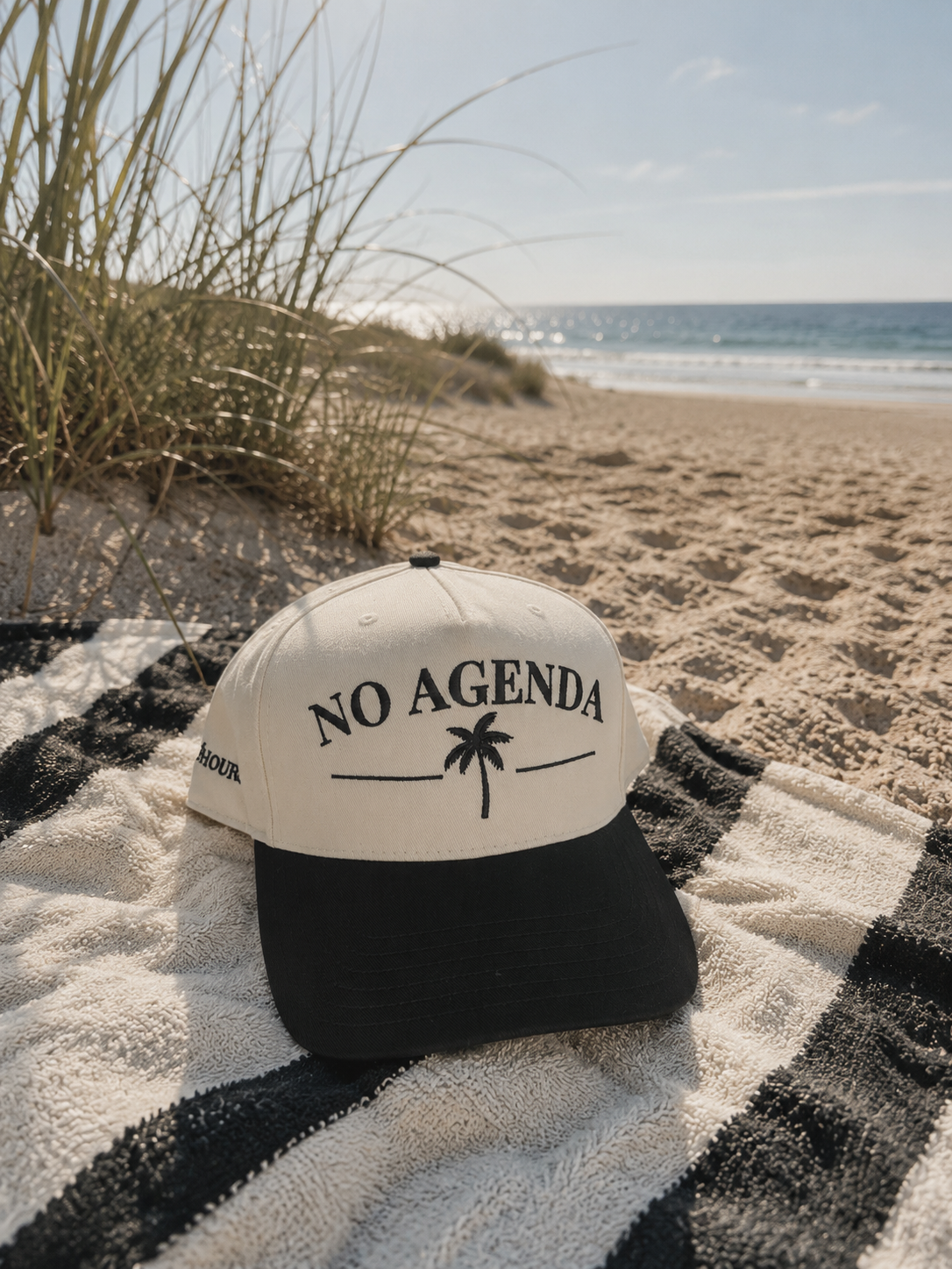 No Agenda black and natural trucker hat by Off Hours sitting on a beach towel on the beach